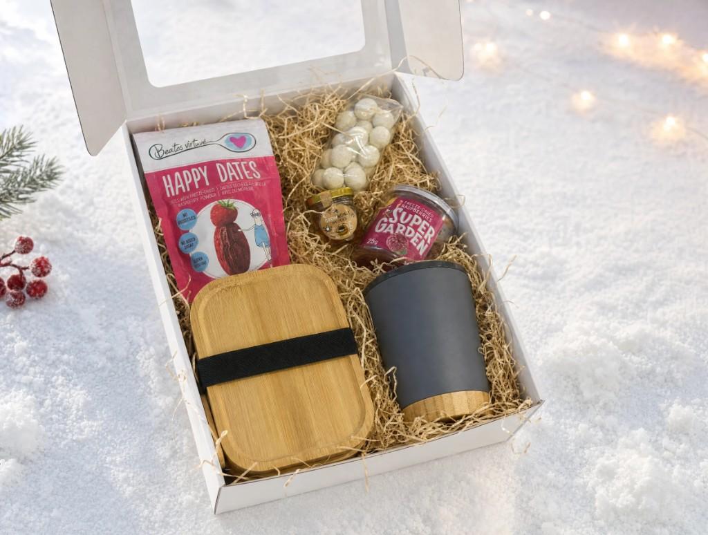 Atvira balta dovanų dėžutė ant sniego: maisto dėžutė, termosinis puodelis, Super Garden avietės, Happy Dates datulės, riešutai ir Grow Honey medus.