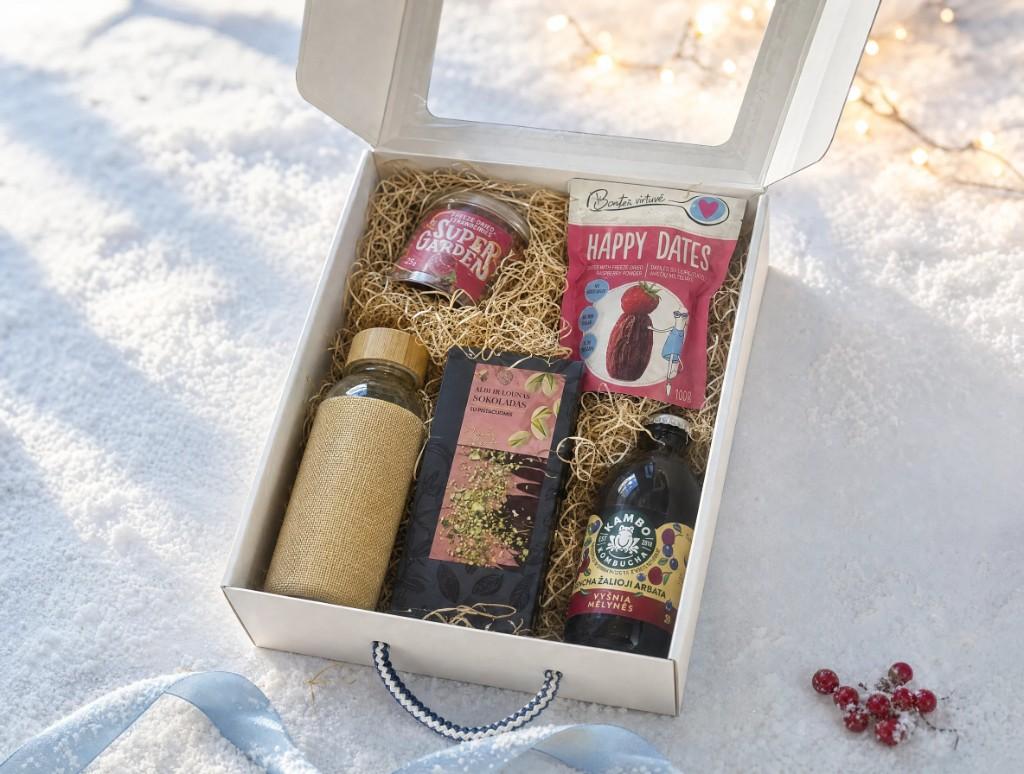 Atvira balta dovanų dėžutė ant sniego: Super Garden indelis, Happy Dates datulės, šokoladas, gertuvė su džiuto įmaute ir Kambo kombučia, šalia žydras kaspinas ir raudonos uogos.